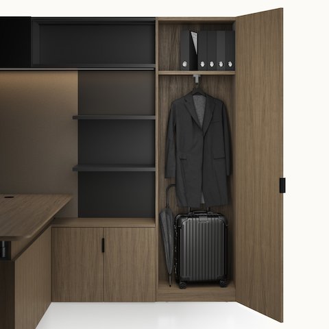 A Geiger One Private Office in Natural Flat Cut Walnut with an open wardrobe containing personal items, a height adjustable desk, and an overhead LED light.