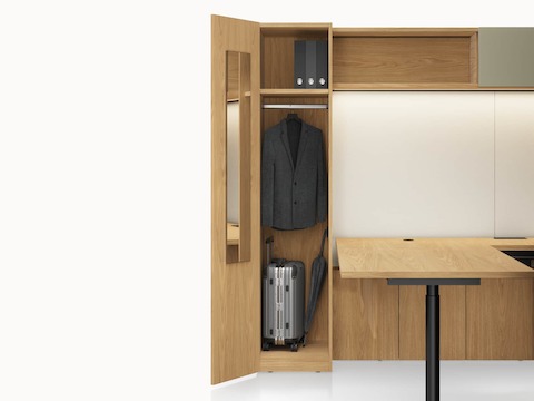 A Geiger One Private Office in Natural Flat Cut Oak with an open wardrobe containing personal items, a height adjustable desk, and an overhead LED light.